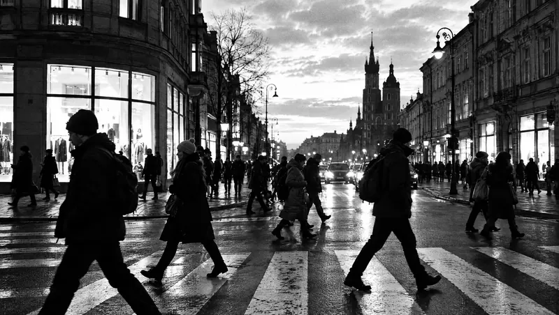 Czarno-białe zdjęcie ruchliwego przejścia dla pieszych o zmierzchu: jasne witryny, niebo i ciemne sylwetki przechodniów